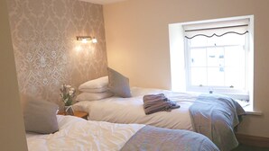 A bedroom with two beds, a window with a view, a nightstand with a vase of flowers, and a wall-mounted light fixture. - Manor House Inn (Haltwhistle)