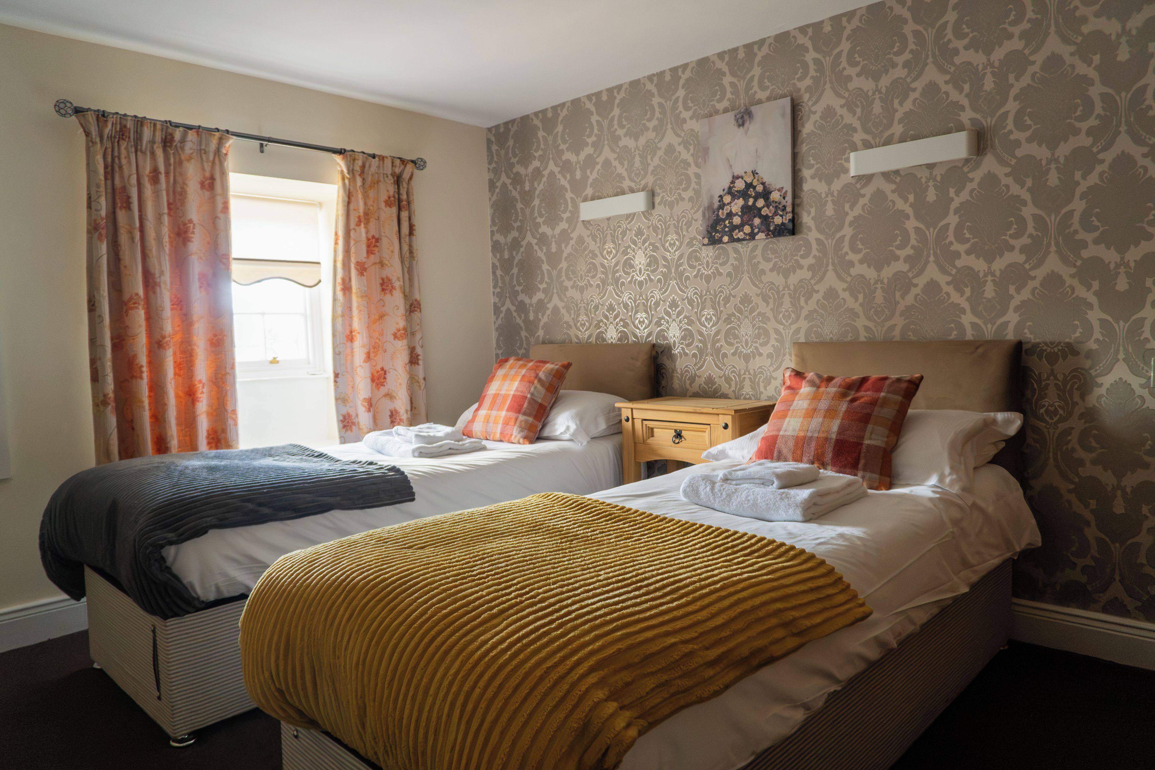 A double bed with a yellow throw blanket, a bedside table with a lamp, and a window with curtains.