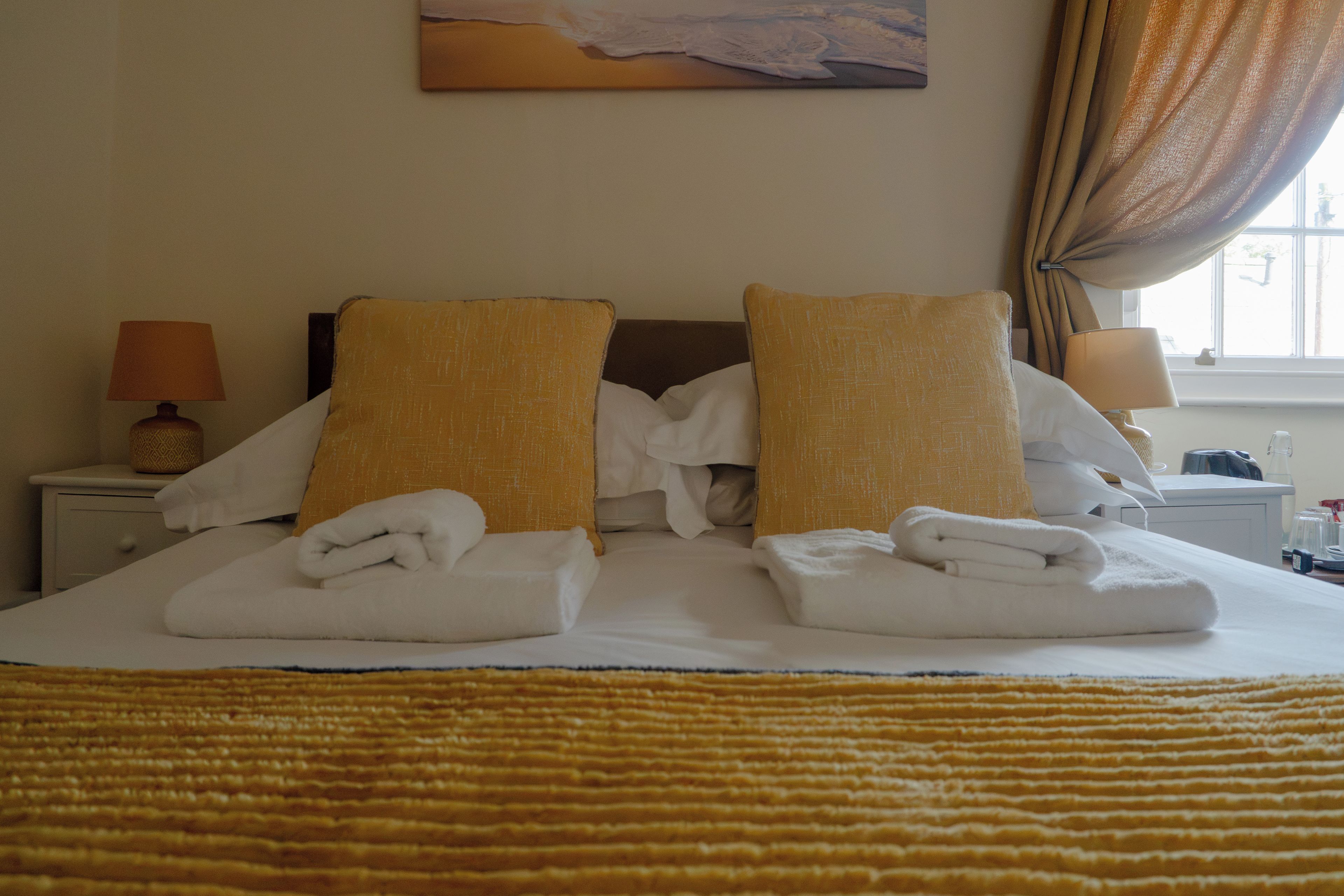A hotel room with a bed, pillows, a nightstand, a lamp, and a window with curtains.