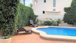 A swimming pool with lounge chairs, surrounded by a garden with various plants and a building in the background. - Bright Villa 20mtrs from the sea (Comunidad Valenciana)