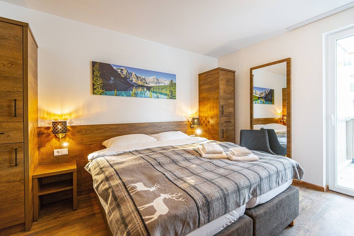 A bedroom with a bed, wooden wardrobe, bedside lamps, a painting, and a mirror.