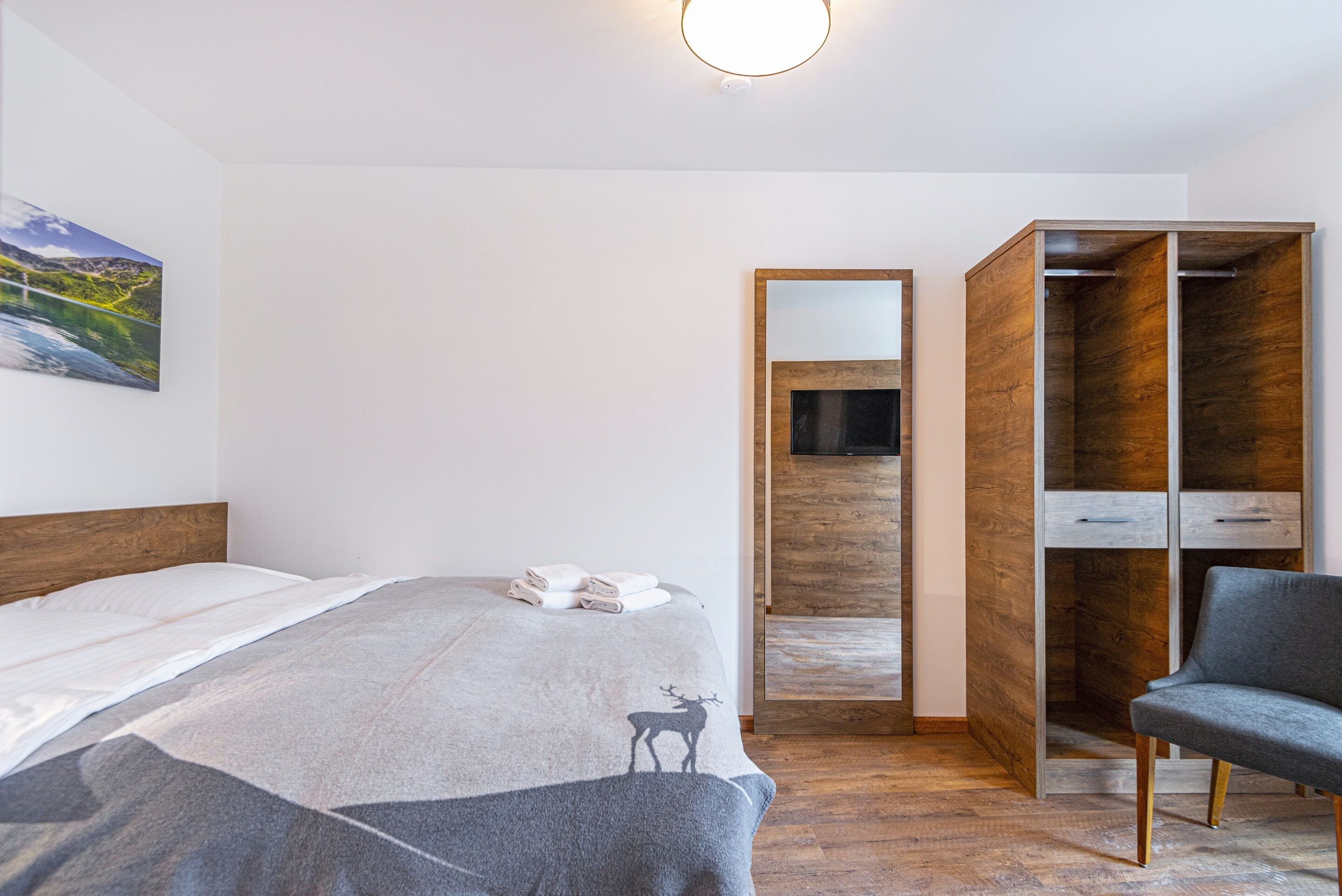 A bedroom with a bed, a gray blanket with a deer design, a gray chair, a wooden wardrobe, and a painting on the wall.
