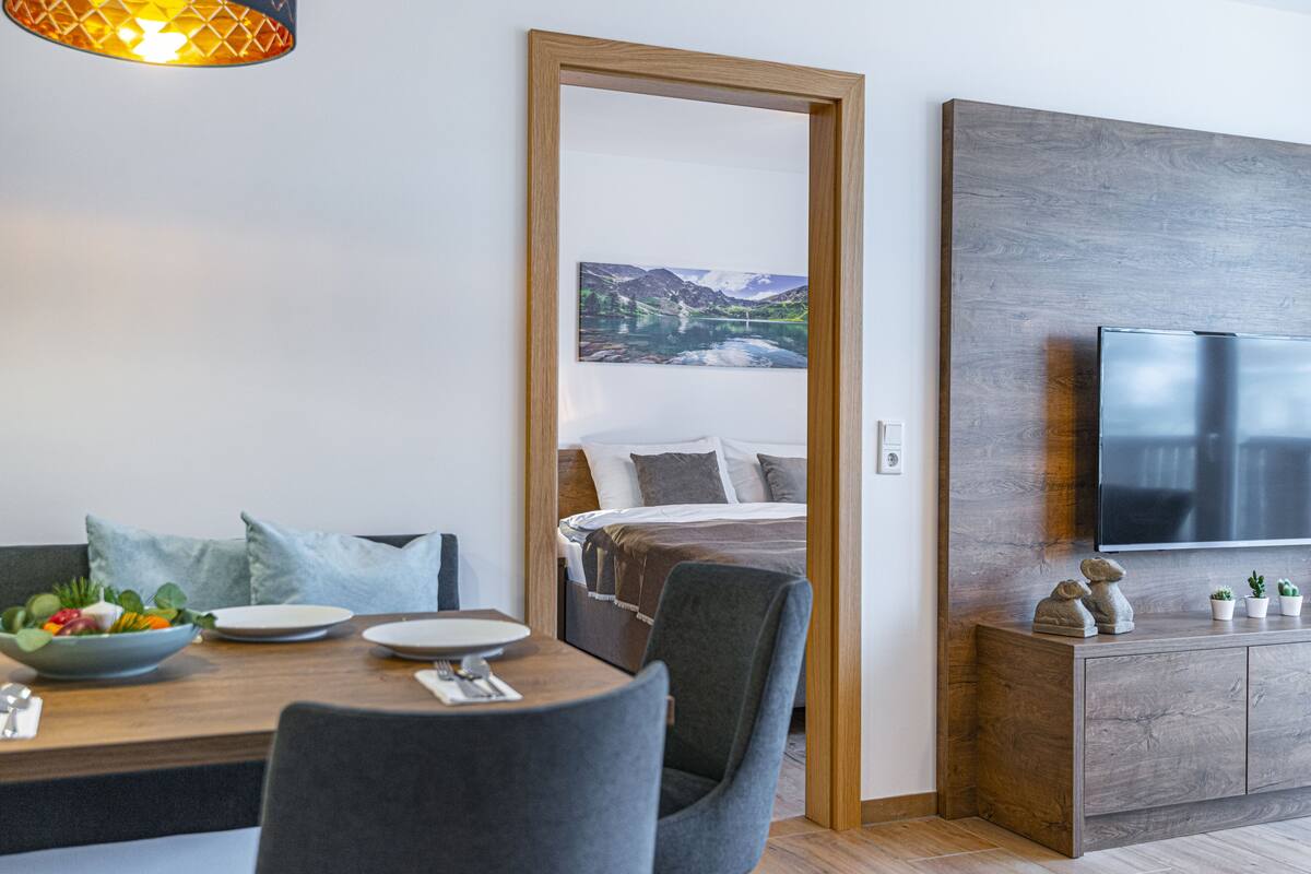 A modern hotel room with a wooden dining table, a bed with a mountain view, a TV on a wooden cabinet, and a small plant on the cabinet.