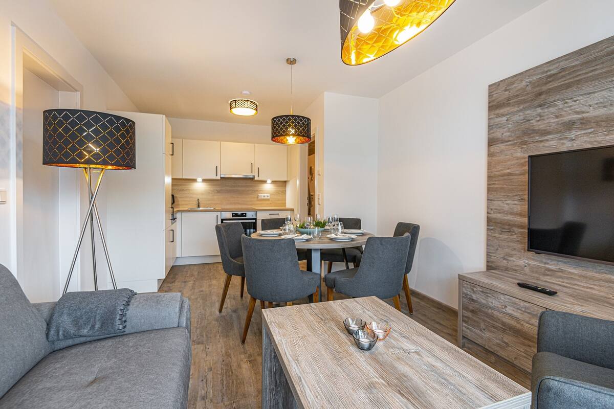 A modern living room with a dining area, grey sofas, a wooden coffee table, and a TV mounted on a wooden wall.