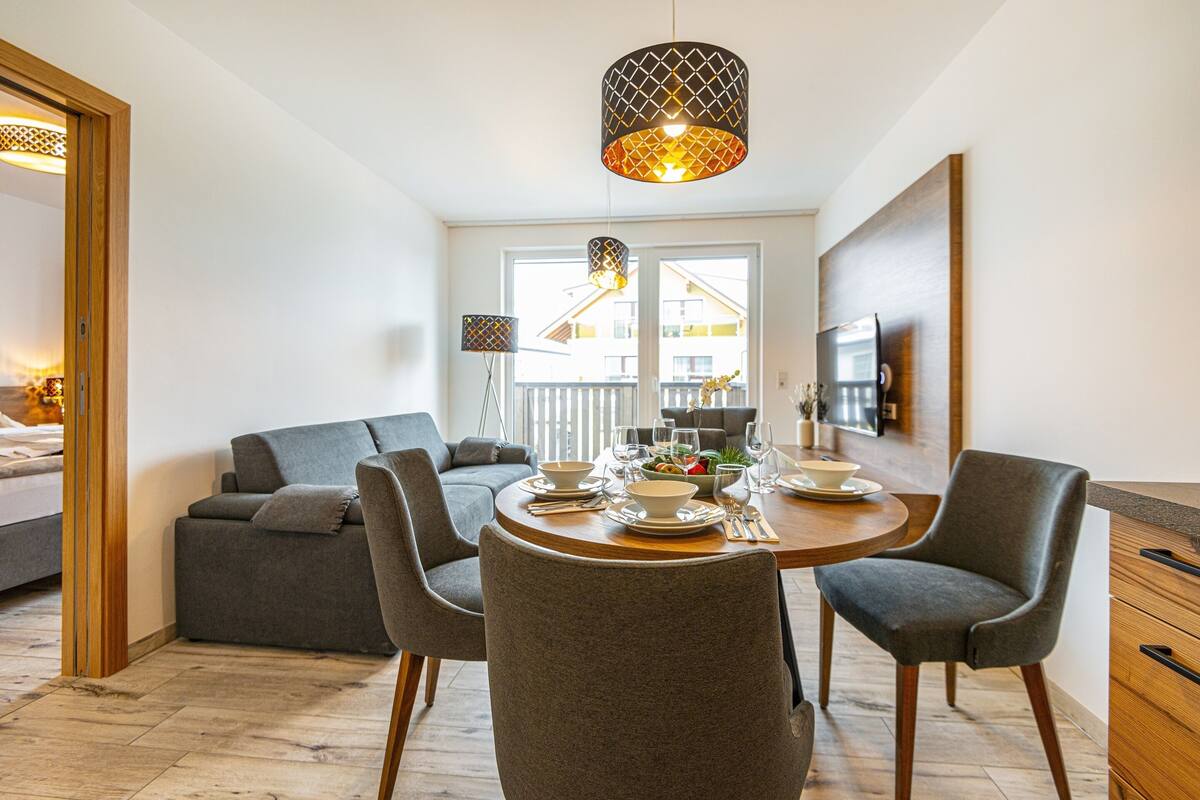 A dining area with a round table set for four, surrounded by grey chairs. There is a sofa and a TV mounted on the wall.