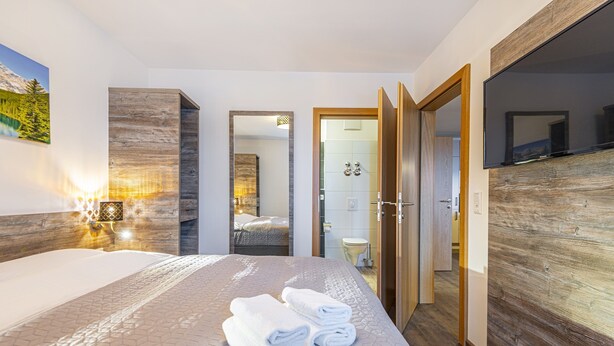 A modern hotel room with a large bed, a wooden headboard, a mounted TV, and a bathroom visible through an open door. - Haus Bergblick (Bad Hofgastein)
