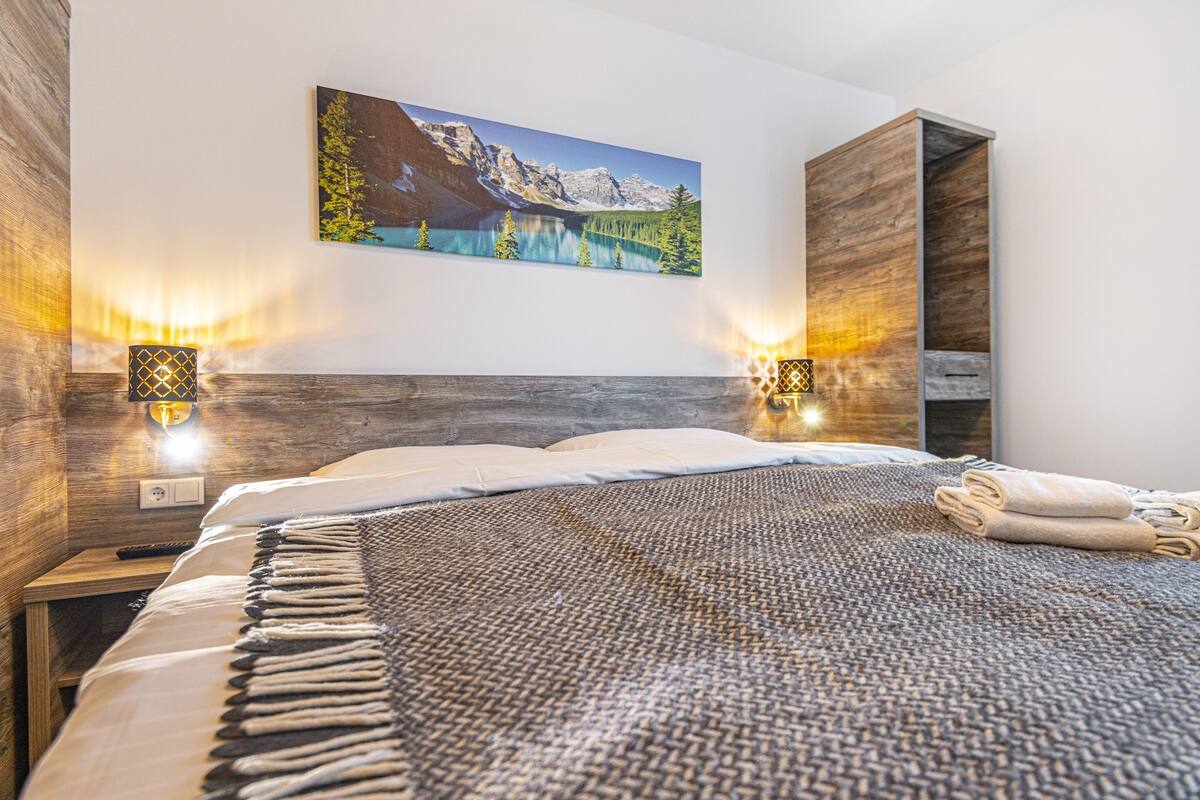 A bedroom with a large bed, wooden headboard, two wall-mounted lamps, a painting of a mountain landscape, and a wooden wardrobe.