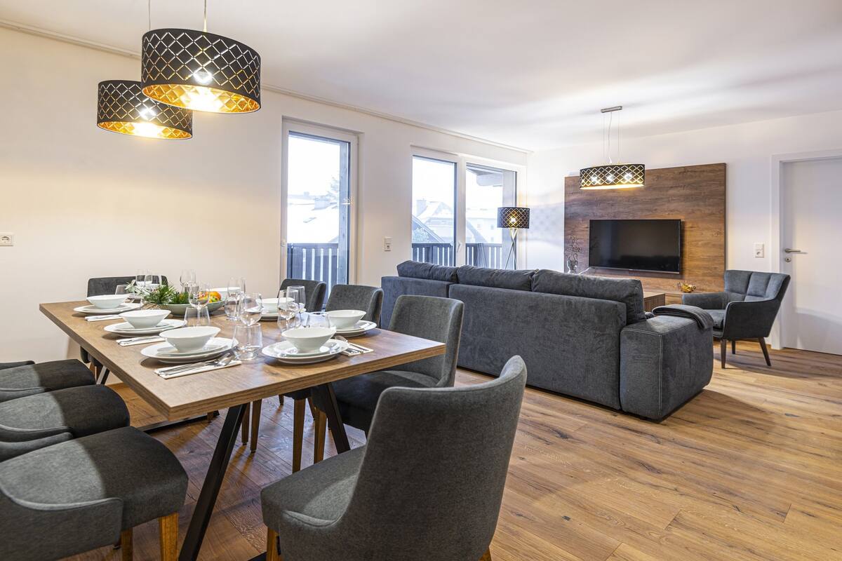 A modern living room with a dining area, grey sofas, a wooden dining table set for four, and a flat-screen TV mounted on a wooden wall.