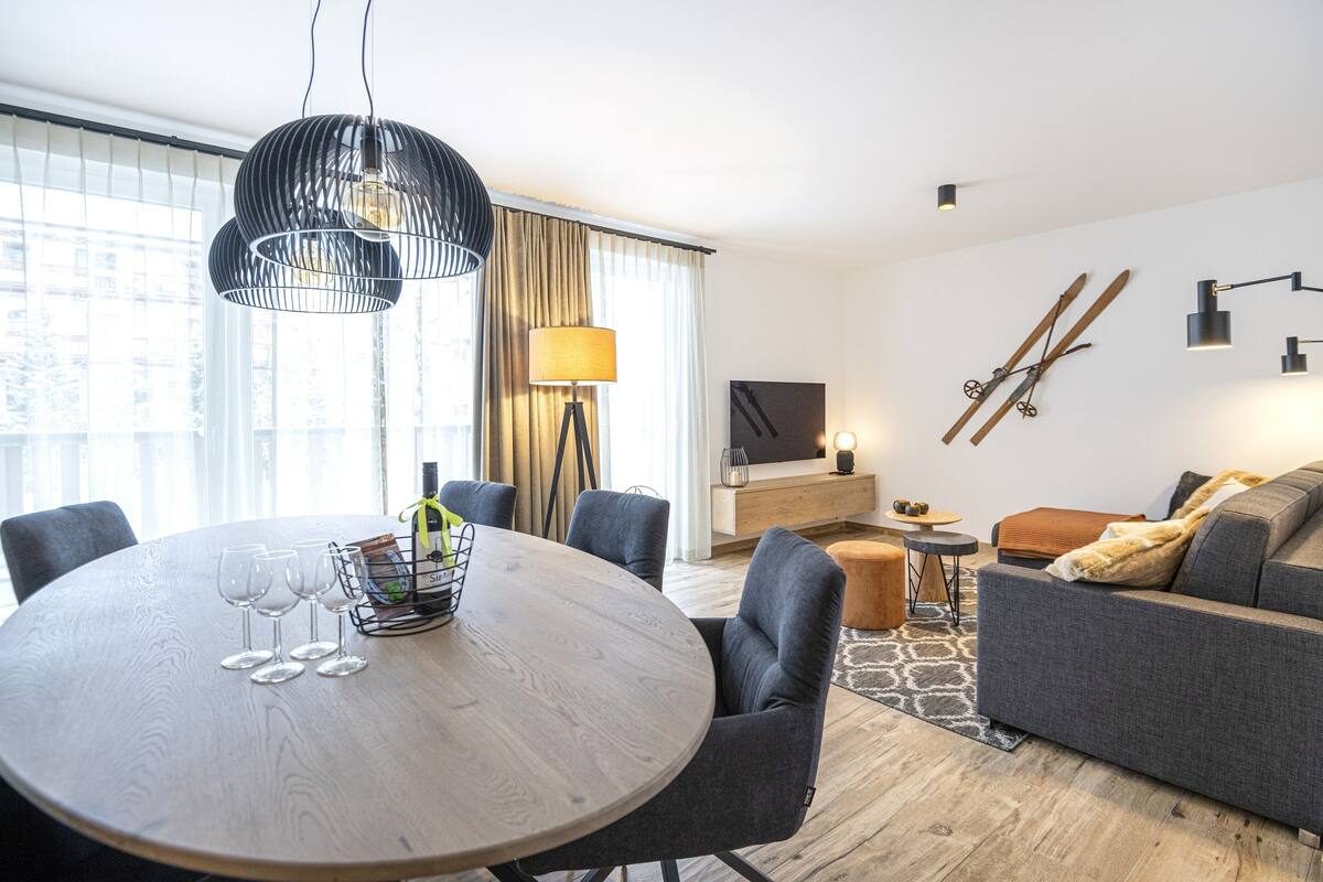 A modern living room with a round wooden table, grey sofas, and a TV mounted on the wall.