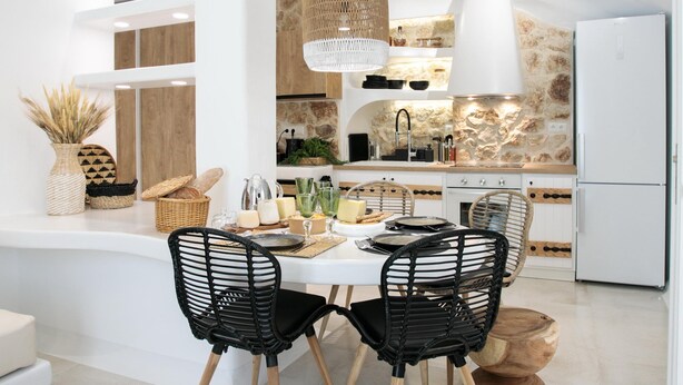 Een moderne keuken met witte kasten, een steen achterwand en een centraal eiland. Er staan twee zwarte stoelen met houten poten, een geweven mand en een geweven vloerkleed.