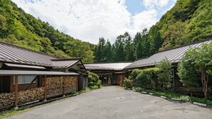 Un edificio japonés tradicional con fachada de madera y tejado de tejas, rodeado de abundante vegetación y un cielo despejado.