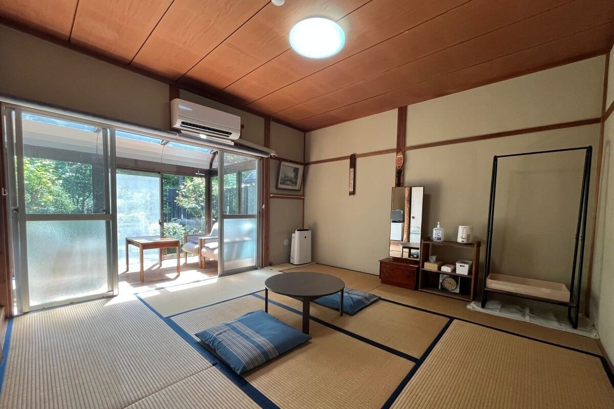 Um quarto japonês tradicional com tatames, uma porta deslizante para uma varanda, uma mesa redonda e um pequeno espaço de estar.