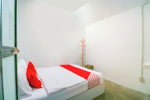 A small, single-bed room with a white bedspread and a red accent pillow. There is a wooden coat rack against the wall and a small wooden stool.