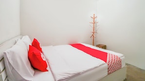 Un llit individual amb roba de llit blanca i un detall vermell, una estant de fusta per a abrics i una petita taula de nit de fusta.