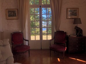 Ein Zimmer mit zwei roten Polstersesseln, einer Holzkommode mit einer Lampe und einem großen Fenster mit Blick auf Bäume und ein Gewässer.