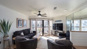 Un salon moderne avec un canapé de couleur foncée, une table basse et un meuble pour la télévision. On y trouve également un ventilateur de plafond, une grande fenêtre donnant sur l’extérieur et une enseigne sur laquelle on peut lire « MAISON DE PLAGE ».