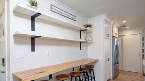 Une cuisine avec des meubles blancs, un comptoir-bar en bois et des tabourets noirs. On y trouve des étagères ornées d’objets décoratifs et une horloge.
