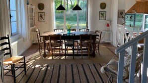 A dining area with a wooden table and chairs, large windows with curtains, and a view of greenery outside.