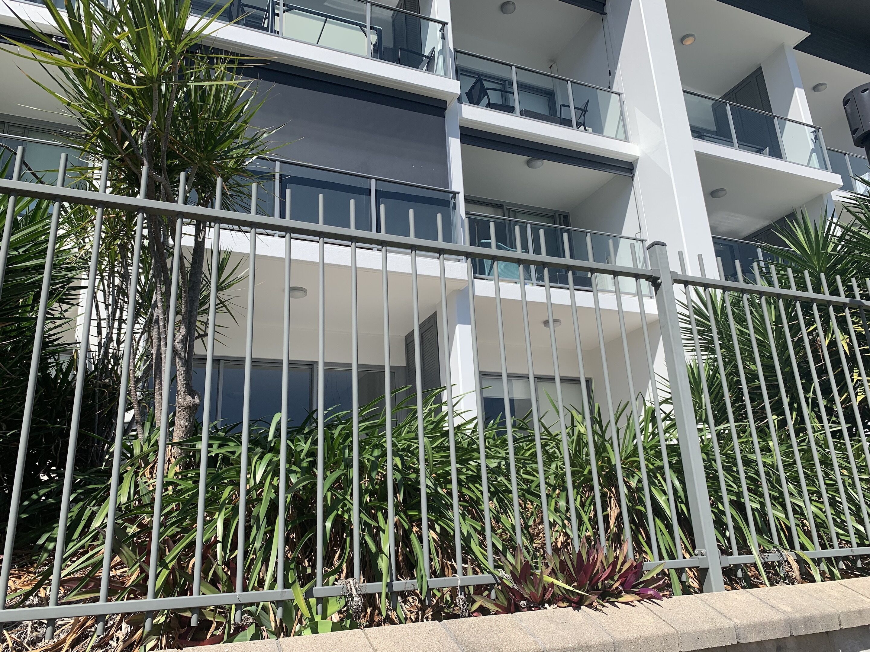 A modern multi-story building with balconies, surrounded by a metal fence and tropical plants.