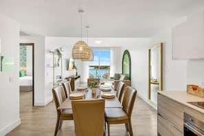 A modern dining area with a large table set for six, surrounded by leather chairs. A hanging light fixture is above the table, and a large window provides a view of the outdoors.