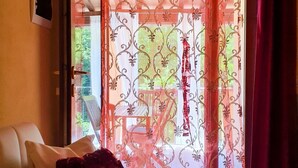 Habitació amb vista per la finestra, cortines decoratives, una cadira blanca i un coixí vermell.