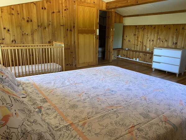 A room with a crib, a bed with floral bedding, a dresser, and wooden walls.