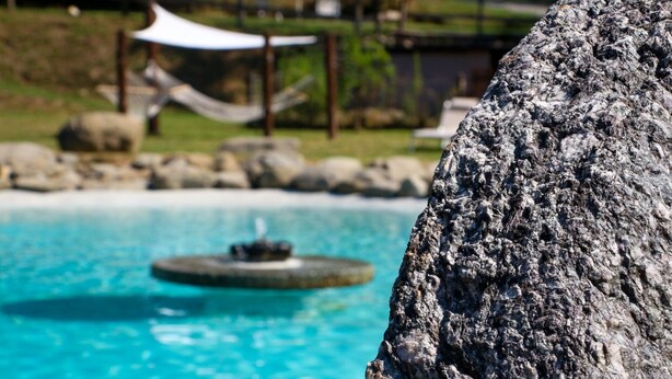 Vista chiara di una piscina con una fontana circolare al centro, circondata da rocce e un'area verde con una struttura a tettoia bianca sullo sfondo.
