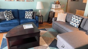 Un salon avec un canapé bleu, un canapé gris, une table basse et un tapis à motifs. Trois tableaux sont accrochés au mur et représentent un coucher de soleil sur l’océan.