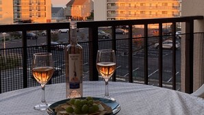 Une table avec une bouteille de Hampton Water, deux verres de vin et une assiette de crackers et de raisins, le tout sur un balcon donnant sur un parking et des immeubles.