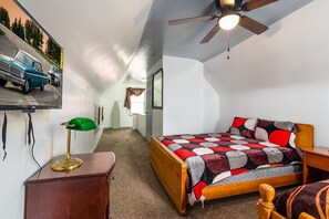 Une petite chambre mansardée, bien éclairée, comprenant un lit, un bureau et un ventilateur de plafond.
