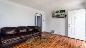 Un salon avec un canapé en cuir marron, une table basse en verre, un parquet en bois, une porte blanche et un téléviseur fixé au mur qui diffuse une scène de voiture.