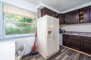 Une cuisine de taille réduite, avec des meubles de rangement en bois foncé, un réfrigérateur blanc et une fenêtre équipée de stores.