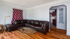 Un salon avec un canapé d’angle en cuir marron, une table basse en verre aux pieds dorés, un rideau rouge à motifs et un plancher en bois.