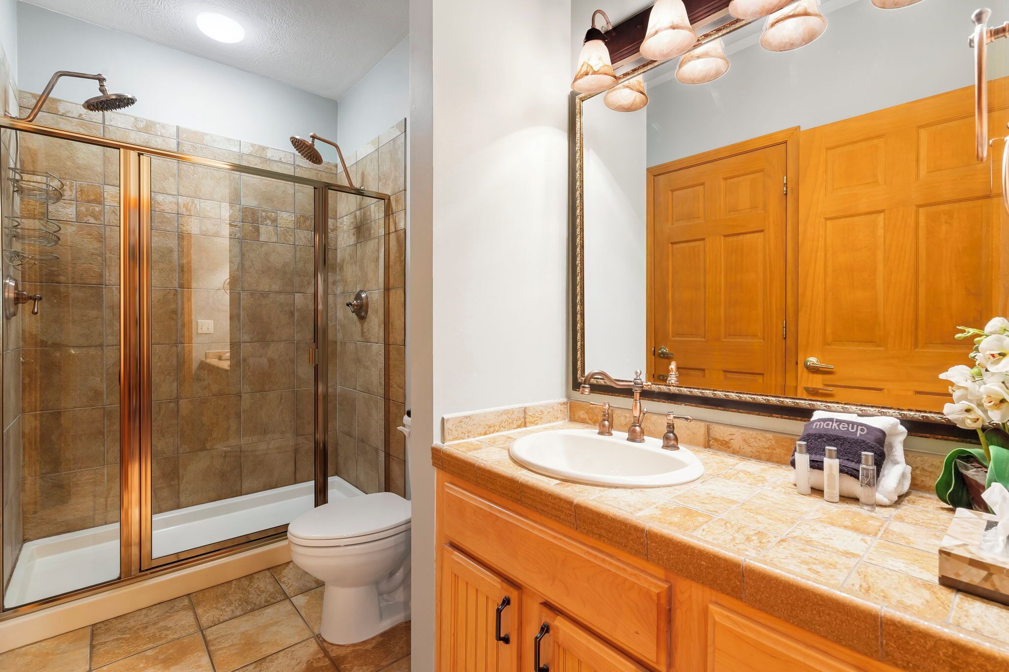A bathroom with a glass-enclosed shower, a white toilet, a vanity with a sink, and a mirror with lights.