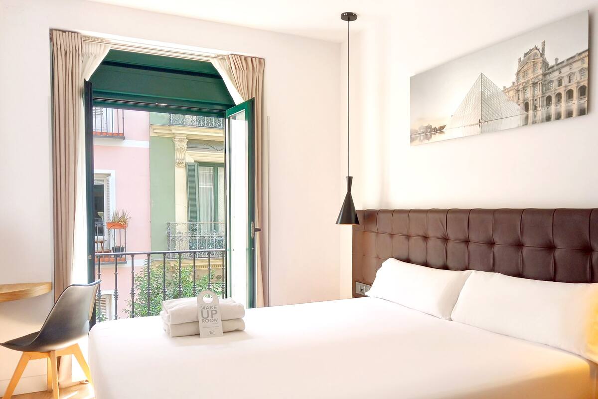 Une chambre d’hôtel avec un grand lit, un balcon donnant sur un immeuble et un tableau accroché au mur.