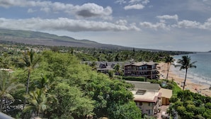 A coastal view with a beach, palm trees, and a large building with multiple balconies overlooking the ocean. - 7th Floor Oceanfront Condo at Mana Kai Resort (Kihei)