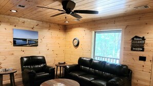 A cozy cabin interior with a leather sofa, two armchairs, a coffee table, a clock, and a window with a view of the outdoors. - "TWO MUCH FUN"   (De Queen)