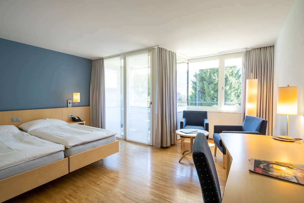 A hotel room with a bed, a desk, a chair, a lamp, and a view of greenery through the window.
