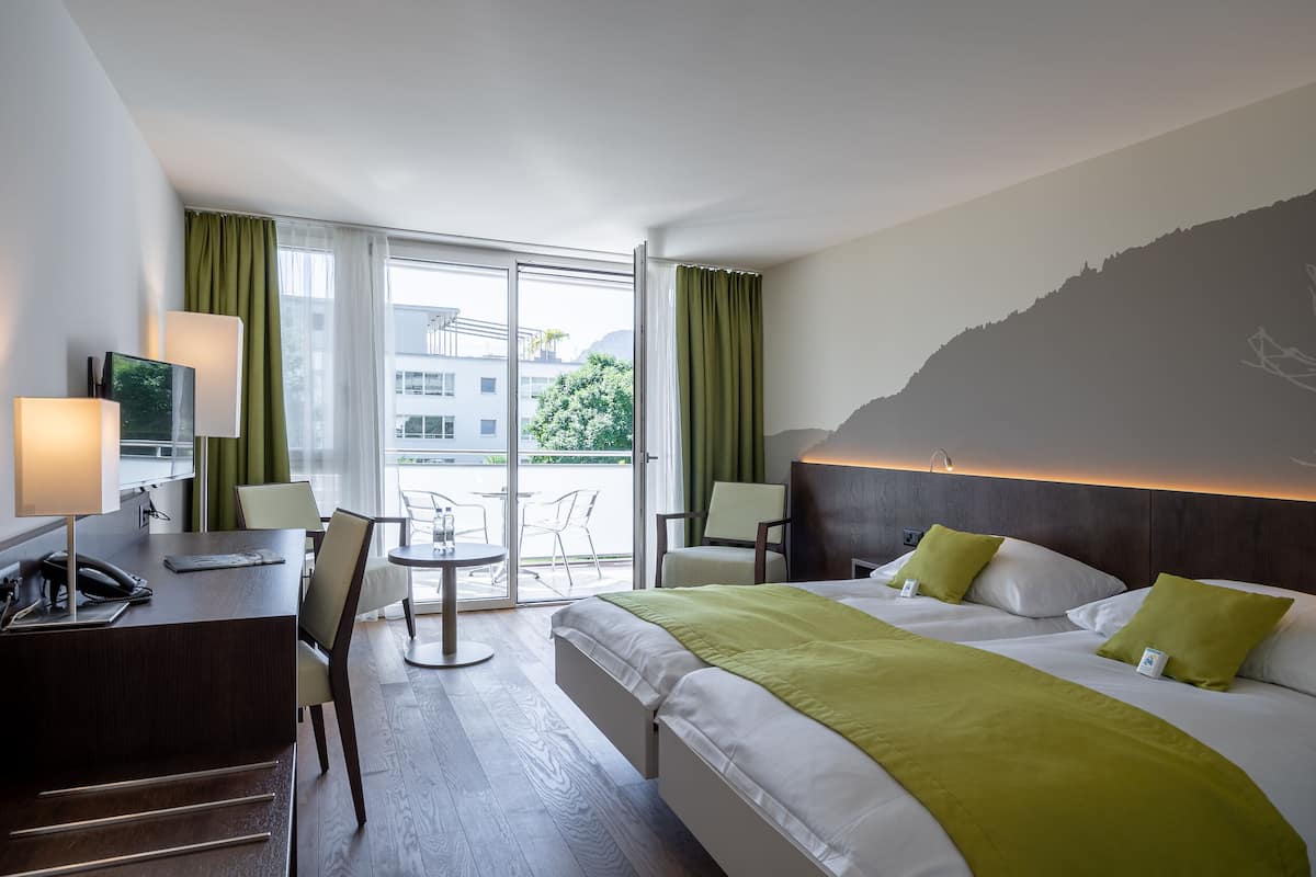 A hotel room with a bed, desk, chairs, and a view of the outdoors.