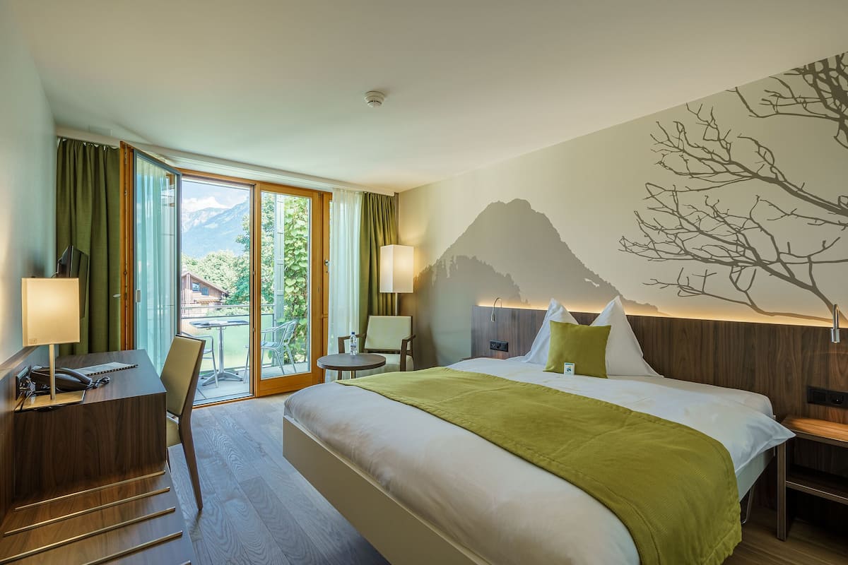 A hotel room with a bed, desk, chair, and a view of mountains through the window.