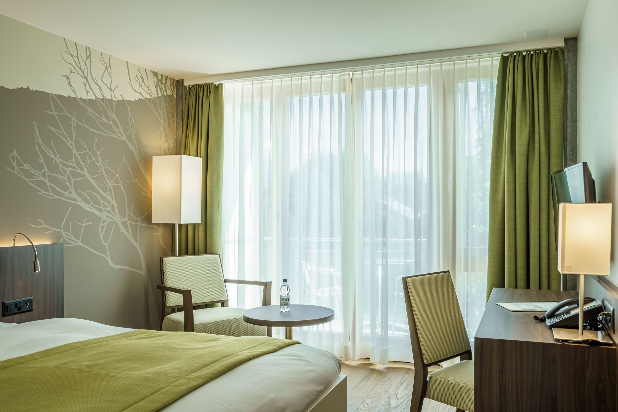 Une chambre d’hôtel comprenant un lit, deux chaises, un bureau avec un téléphone, une lampe et une petite table avec une bouteille.