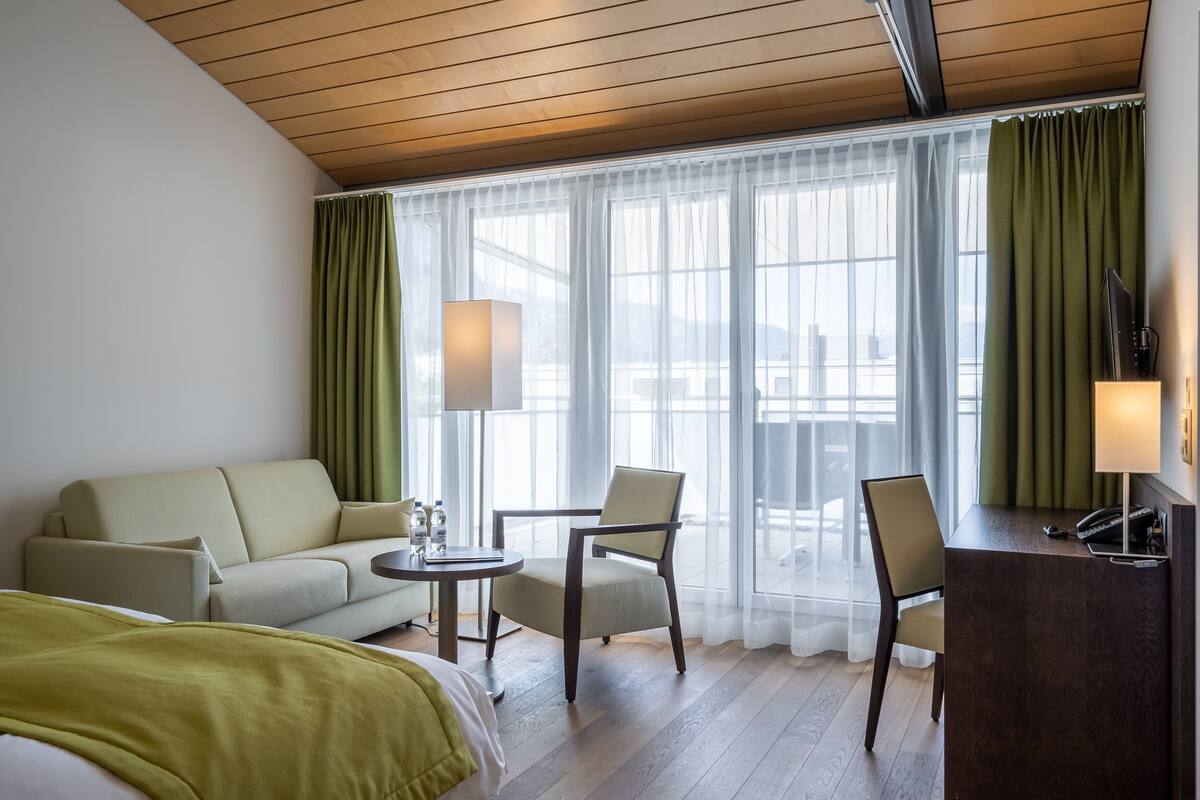 A hotel room with a bed, a sofa, a desk, and a chair. There is a window with curtains, a lamp, and a view of the outside.