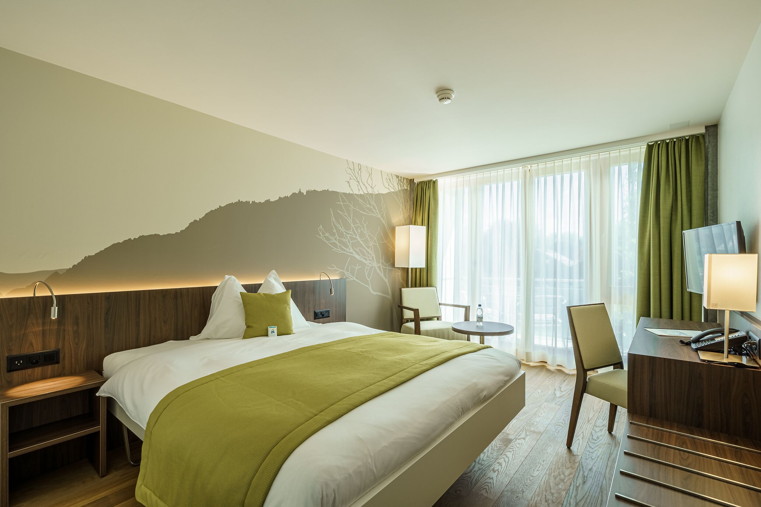 Ett hotellrum med en stor säng, ett skrivbord, en stol, ett litet bord och utsikt över omgivningarna genom fönstret.
