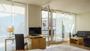 Una habitación de hotel moderna con una cama grande, un escritorio con televisor, un sofá y un balcón con vistas a edificios y montañas.