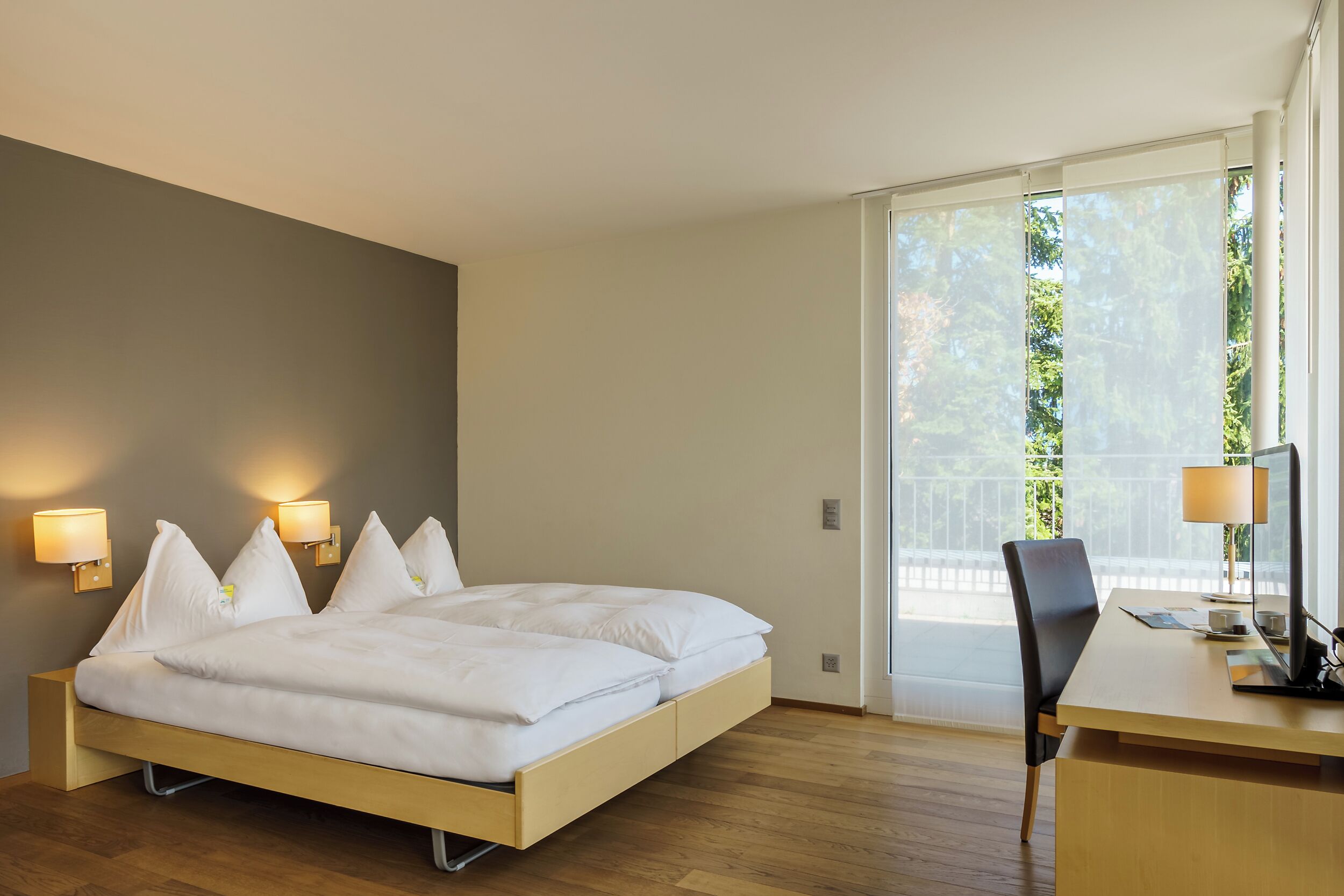 Habitación de hotel moderna con cama, lámparas de noche, escritorio y vistas a zonas verdes a través de amplios ventanales.