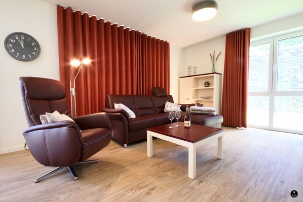 A modern living room with a brown leather sofa, a brown leather armchair, a white coffee table, and a side table. There is a clock on the wall, a floor lamp, and a bookshelf with decorative items.
