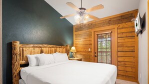 Un dormitorio con cabecera de madera, cama con ropa blanca, mesita de noche con lámpara, ventilador de techo y puerta de madera.