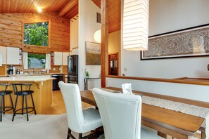 A modern kitchen with wooden accents, a dining area with white chairs, and a large window overlooking a forested area.