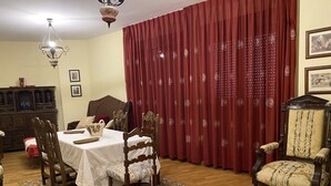 Un comedor con una mesa preparada para cuatro personas, rodeada de sillas de madera. Un candelabro cuelga del techo, y una cortina roja con un diseño circular cubre la ventana.