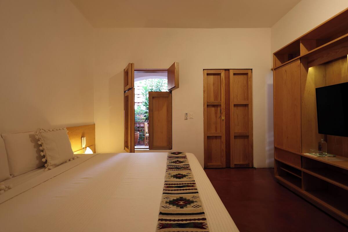 A bedroom with a bed, a wooden wardrobe, a TV, and a window with a view of greenery.